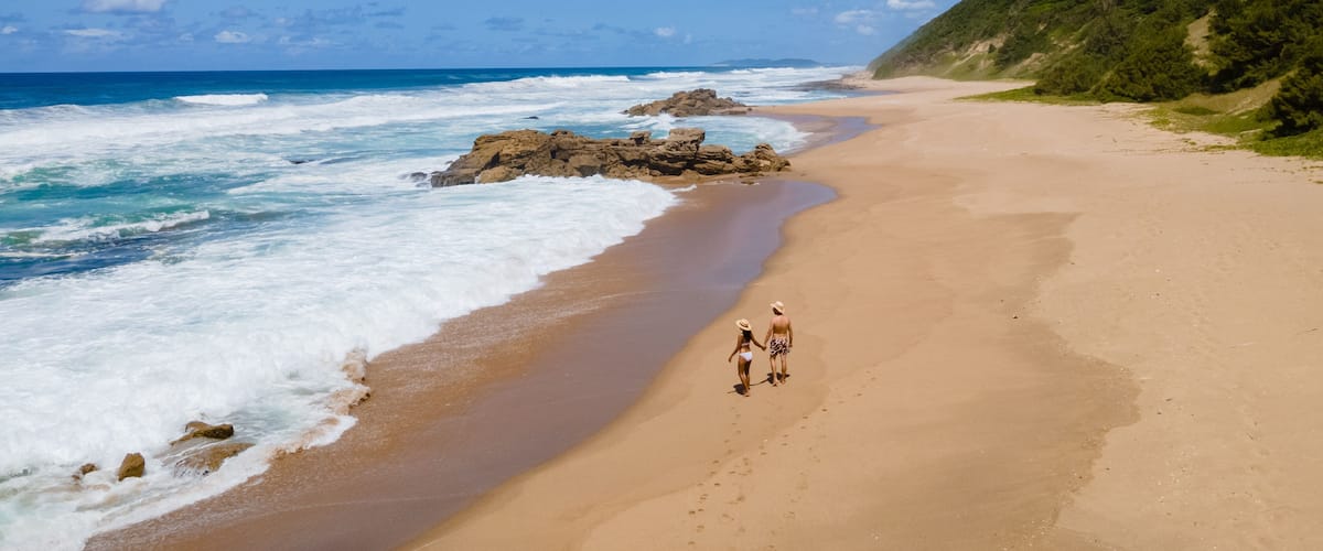 Ein sandiger Strand mit Wellen, ein Paar spaziert am Strand entlang, und ein grüner Hügel ist im Hintergrund zu sehen.