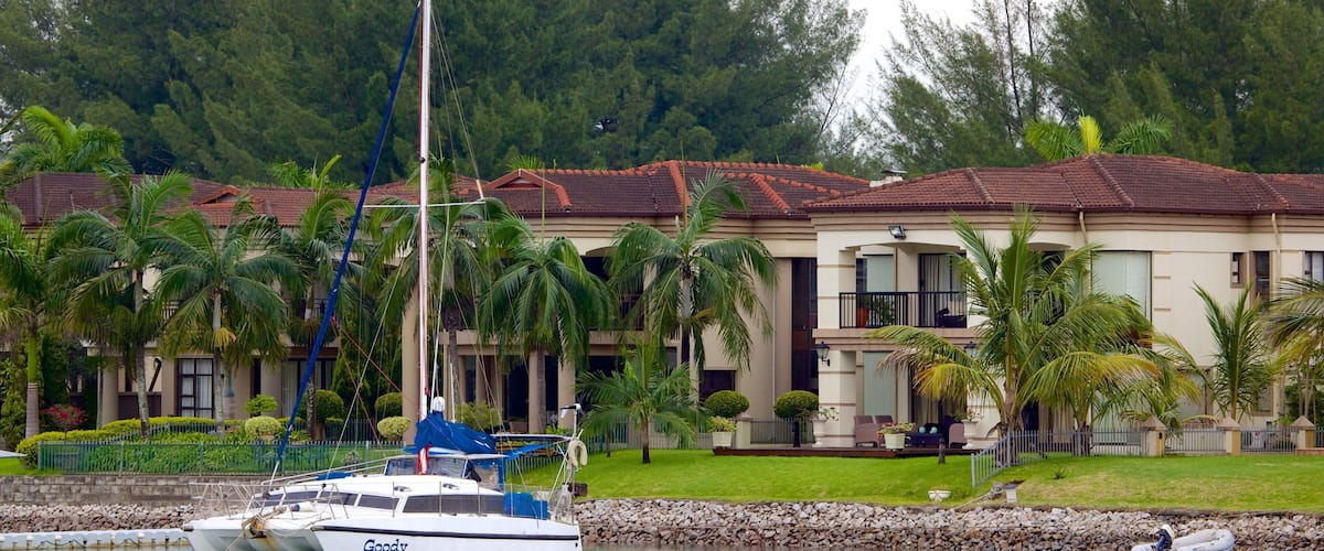 A Goody boat docked near a house with a red-tiled roof, surrounded by palm trees and a rocky shoreline.
