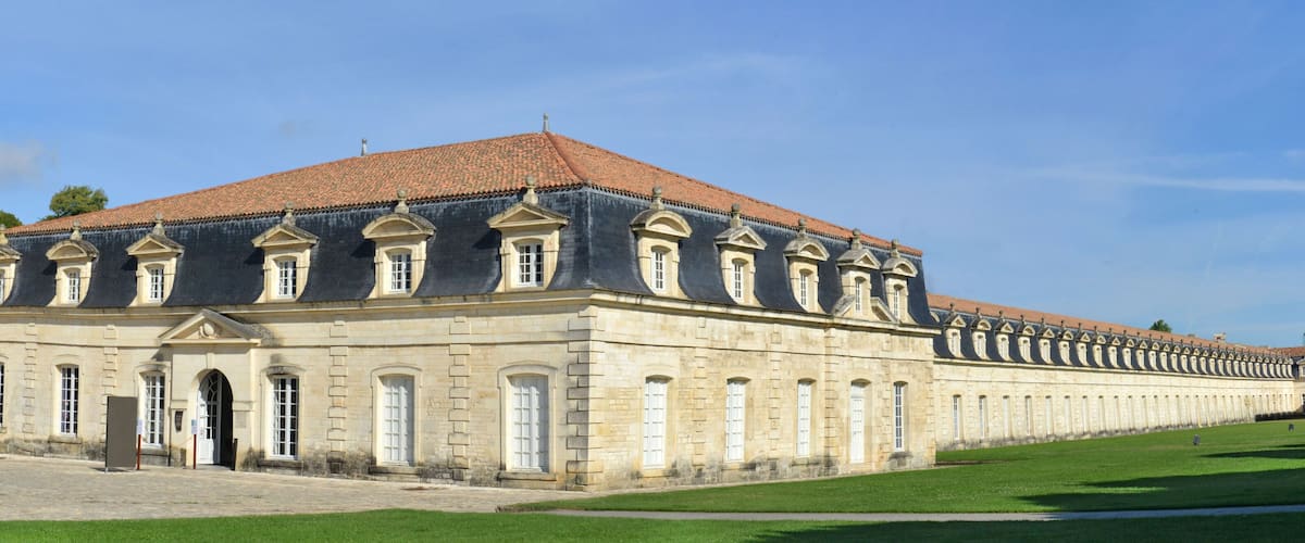 A large, historic building with a long facade, a red-tiled roof, and a well-maintained lawn in front.
