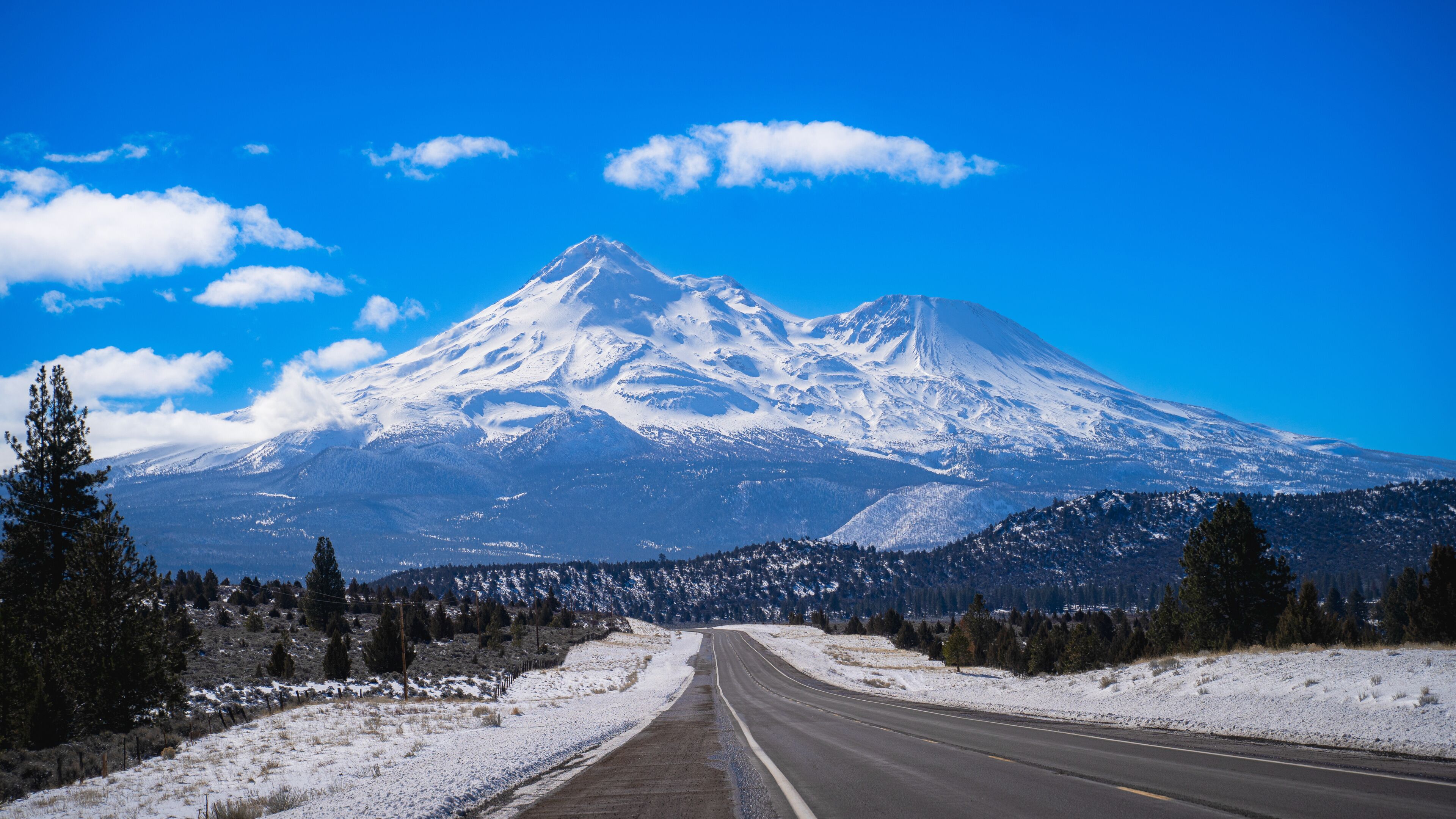 Montanha coberta de neve com céu claro, estrada que a leva e árvores esparsas.