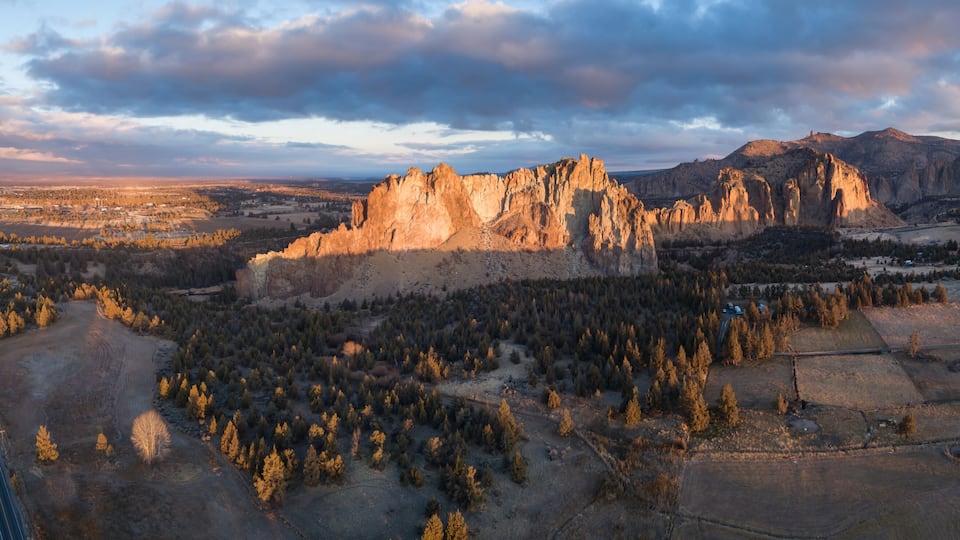 A mountainous landscape with a prominent rocky peak, a dense forested area, and a winding road.