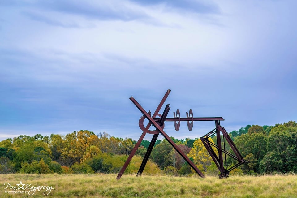 Eine Metallskulptur in einem grasbewachsen Feld, im Hintergrund Bäume.