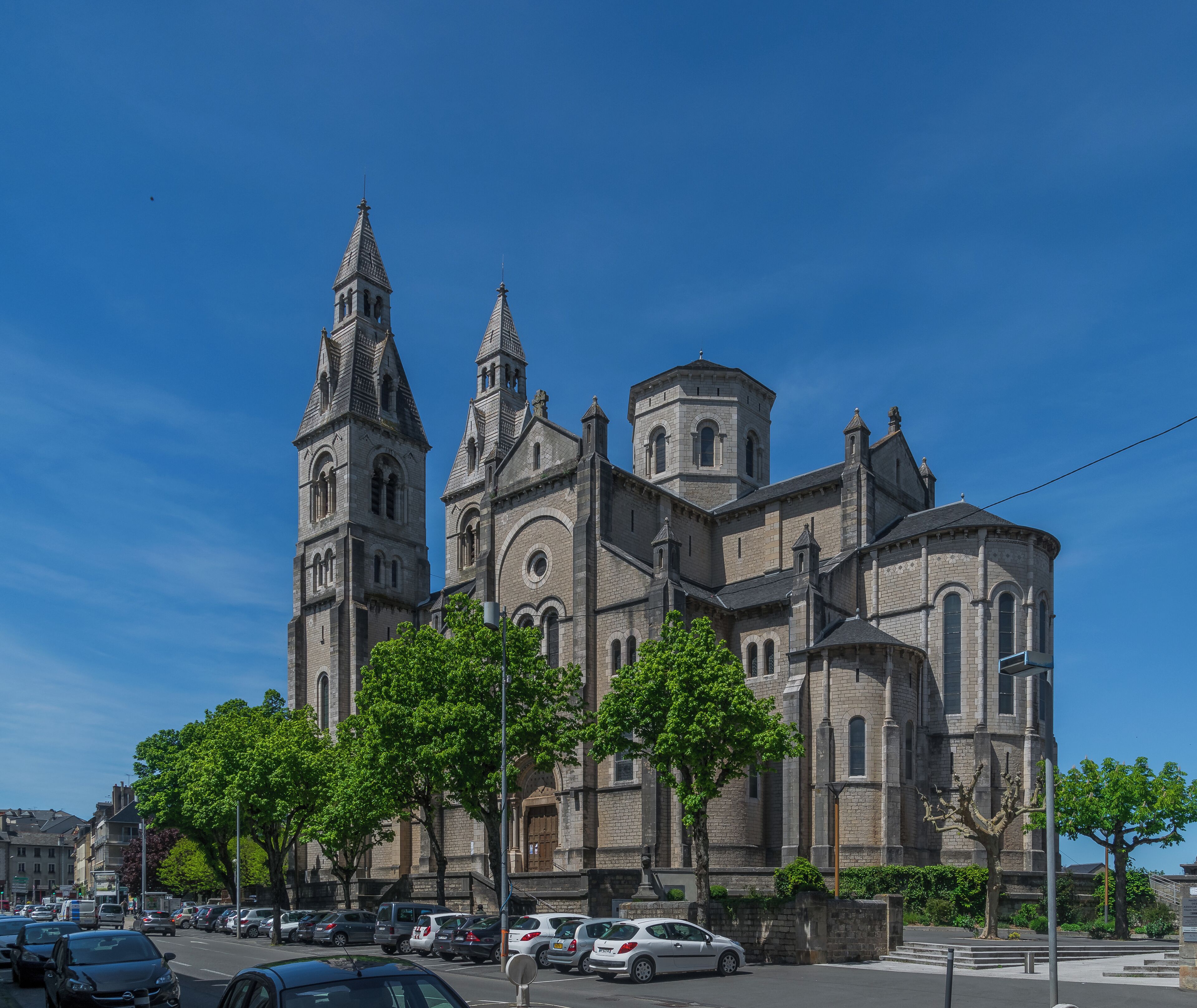 A historic church with two prominent towers and a central dome, surrounded by parked cars and trees.