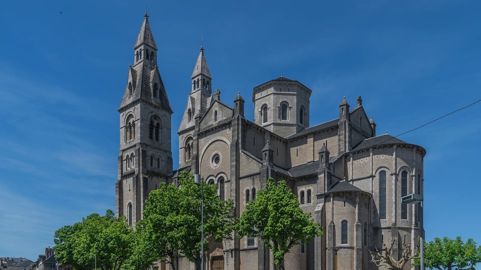 A historic church with two prominent towers and a central dome, surrounded by parked cars and trees.