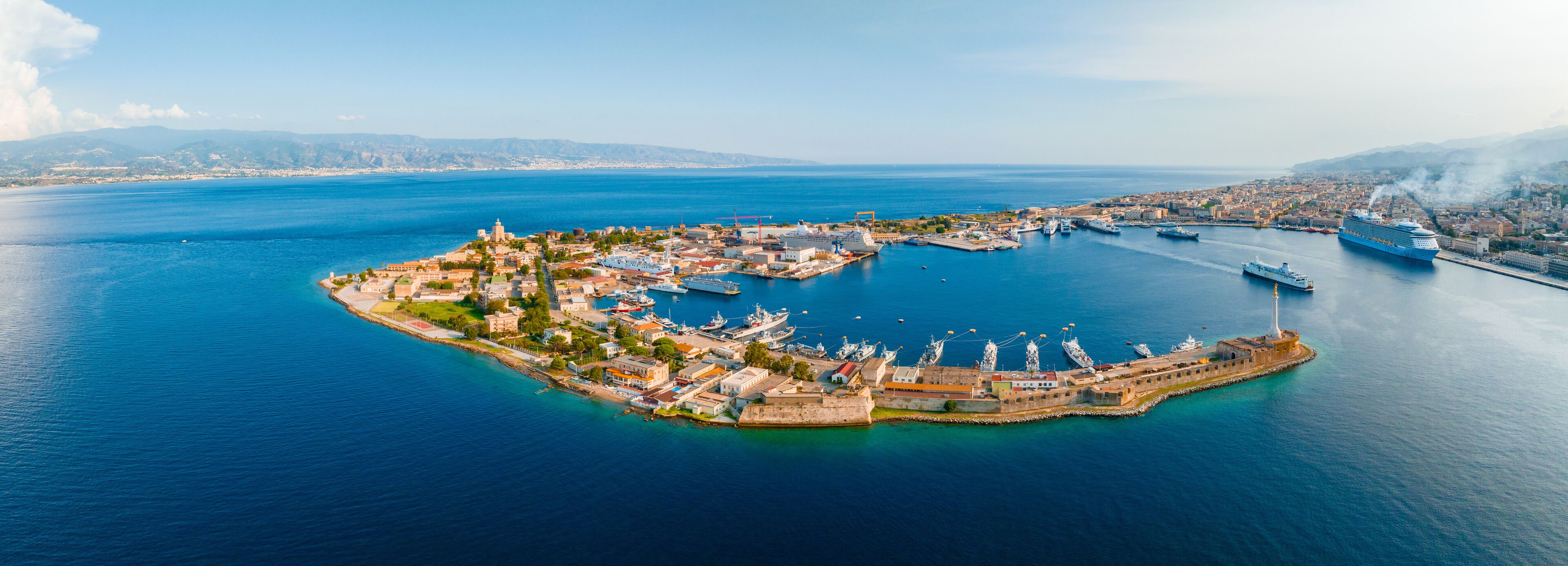 An aerial view of a coastal town with a fortified harbor, surrounded by water and mountains in the background.