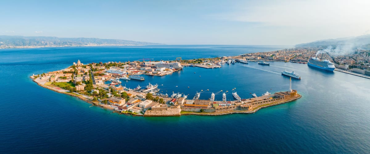An aerial view of a coastal town with a fortified harbor, surrounded by water and mountains in the background.