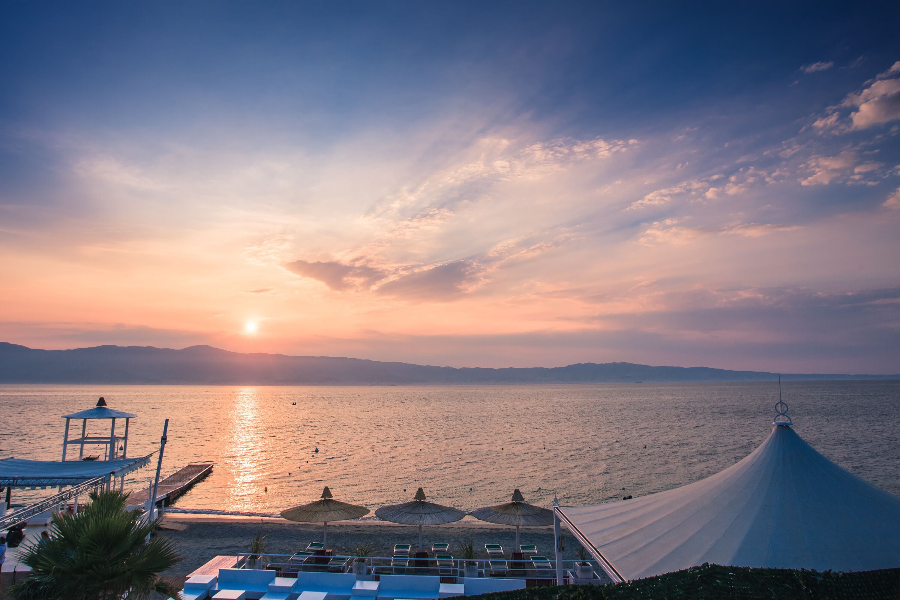 A sunset over a calm sea with a beach, umbrellas, and a boat.