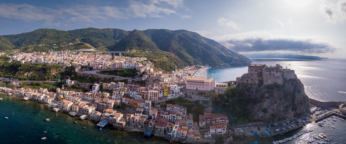 Un pueblo costero con un castillo histórico en un acantilado, rodeado de montañas y un mar azul cristalino.