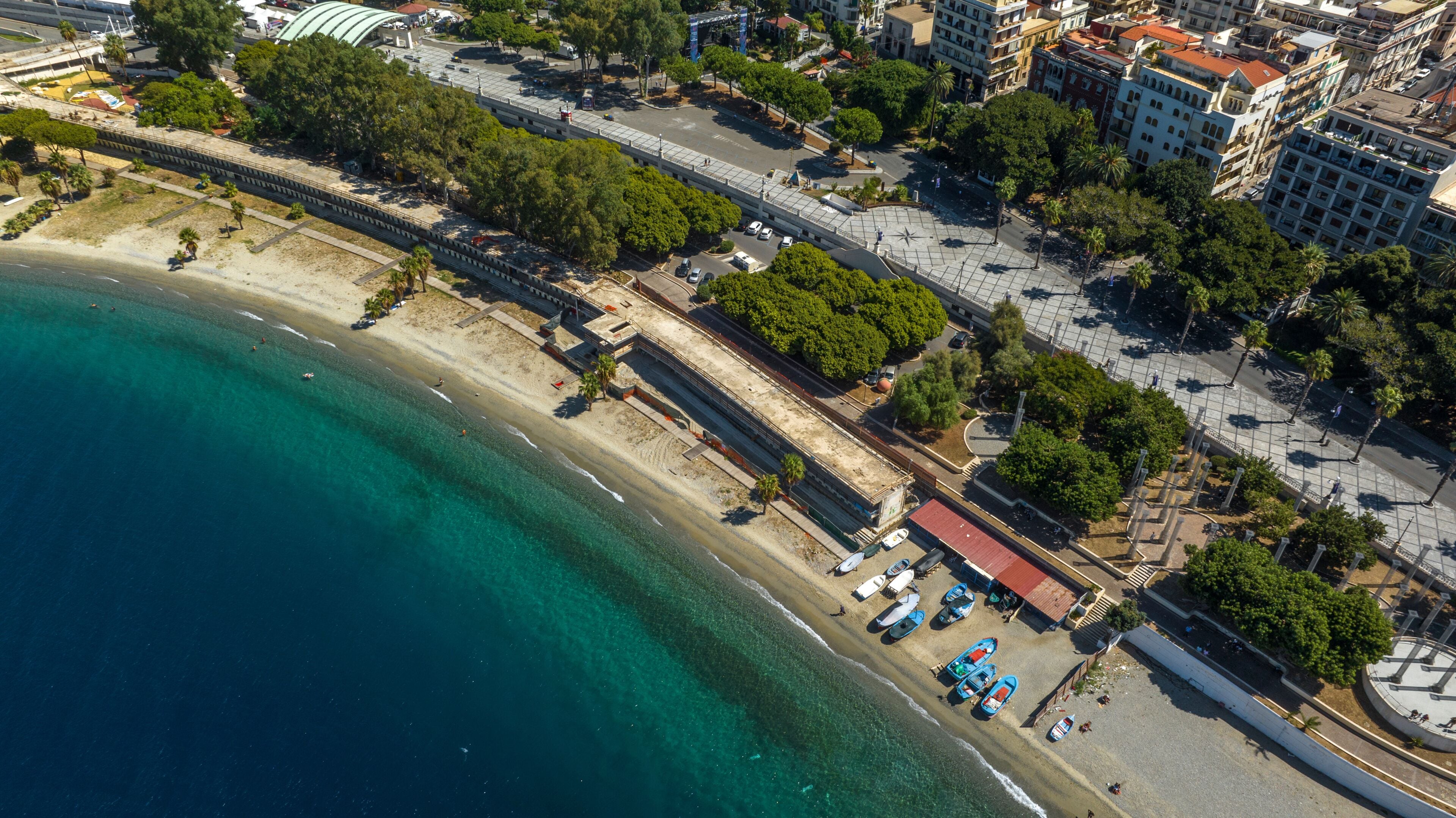 High-angle aerial view of a coastal promenade featuring a grey and white walkway, palm trees, and a urban beach alongside the crystal-clear and blue sea. It is the waterfront of Reggio Calabria, Italy
