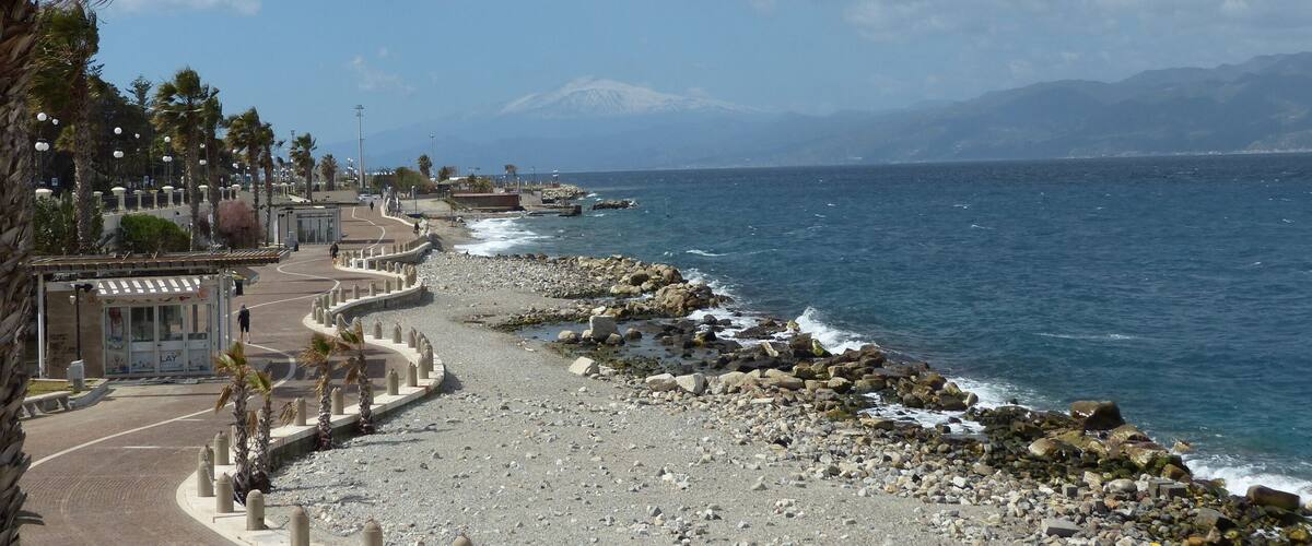 A coastal road with a rocky shoreline, palm trees, and a mountain in the distance.