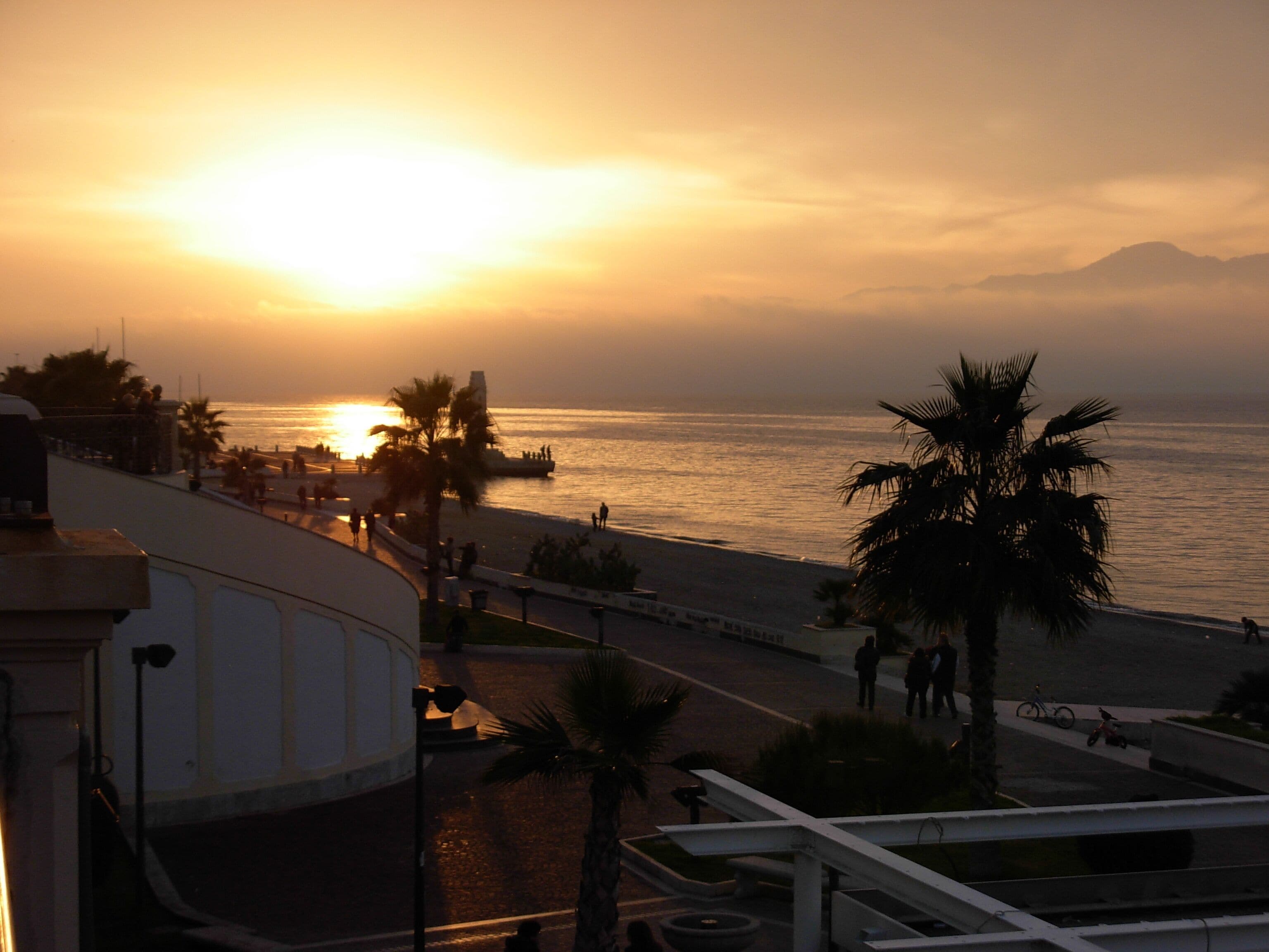 Una puesta de sol sobre una playa con palmeras y un paseo marítimo.