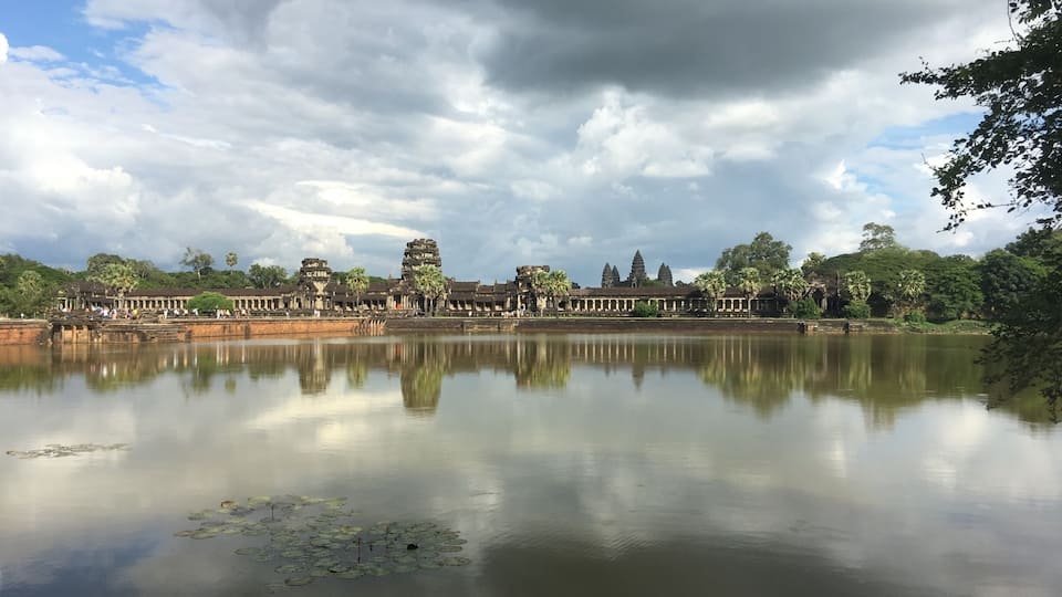 A calm lake reflecting ancient temple structures and lush greenery under a cloudy sky.