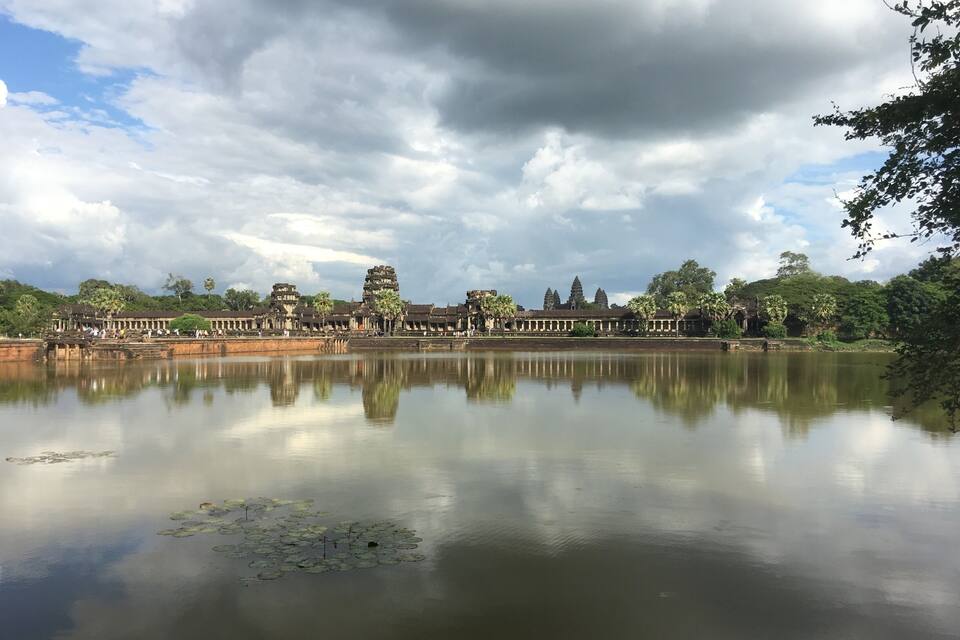 Um lago tranquilo refletindo estruturas de um templo antigo e vegetação exuberante sob um céu nublado.