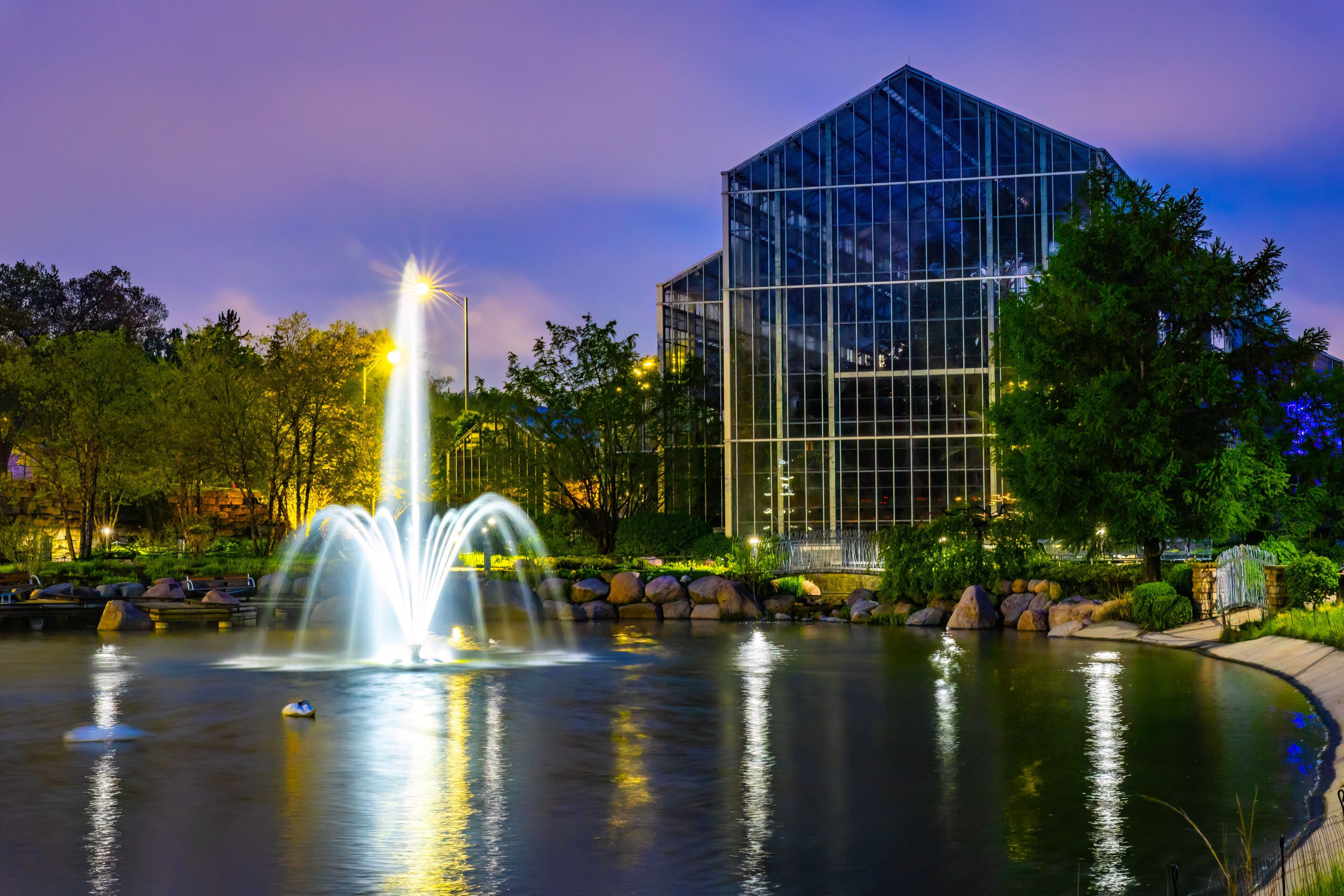 A nighttime scene with a water fountain, a glass building, and surrounding greenery.