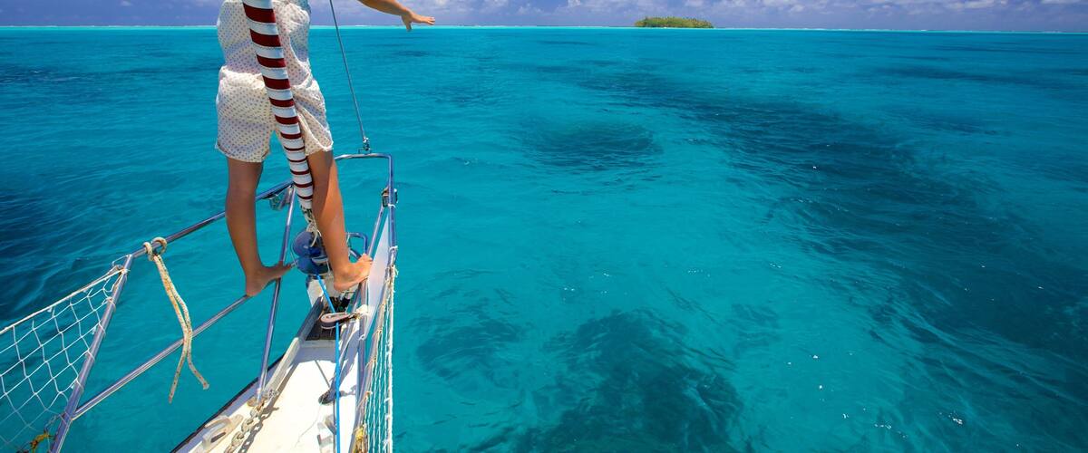 A person on a sailboat in turquoise waters with a clear sky and a small island in the distance.