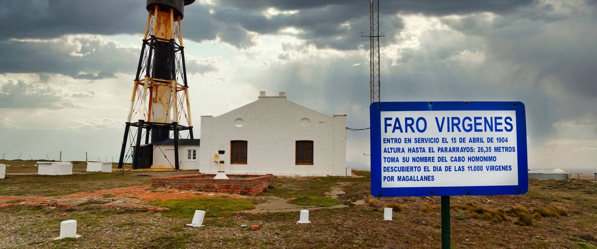 A lighthouse named Faro Virgines with a signboard, a white building, and a cloudy sky.