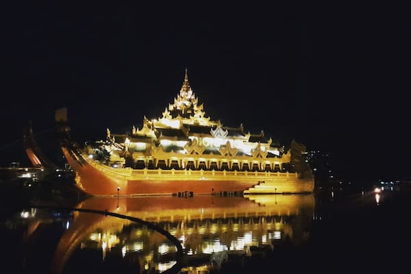 A large, illuminated ship with multiple decks and a traditional design, docked at night with its reflection visible on the water.