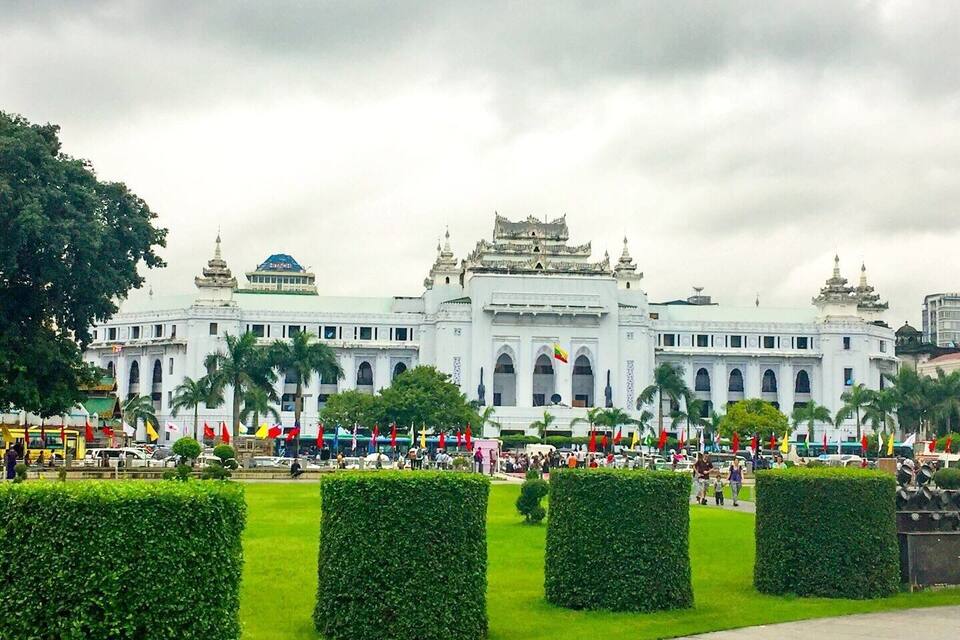 A large white building with traditional architecture, surrounded by green hedges and palm trees.