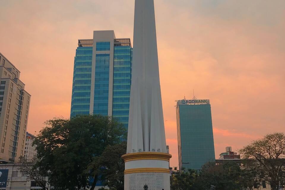 A tall monument with inscriptions, surrounded by a park with people and modern buildings in the background.