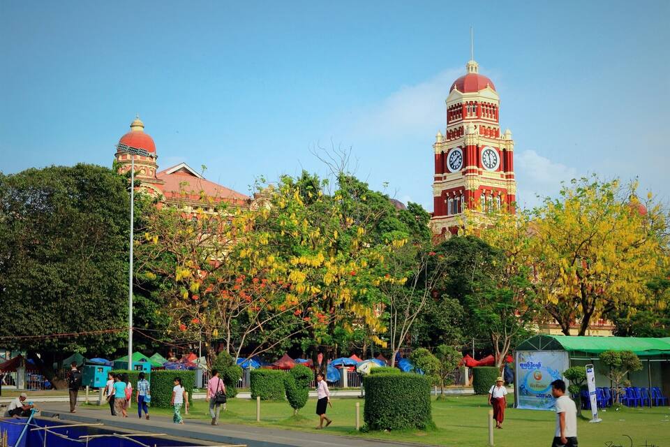 A large clock tower with a red and white facade, surrounded by lush greenery and a clear blue sky.