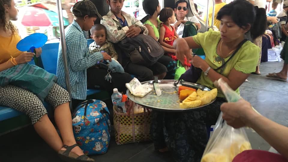 A crowded outdoor market with people sitting and standing, a food vendor serving food, and various bags and items on the ground.