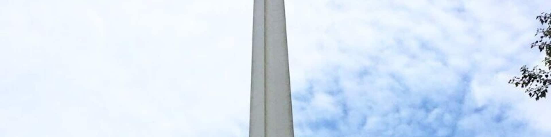 A tall monument in a park with a clock tower in the background.