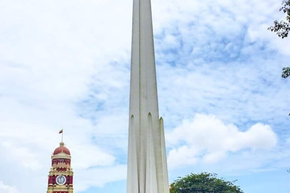 Un monumento alto en un parque, con una torre del reloj al fondo.