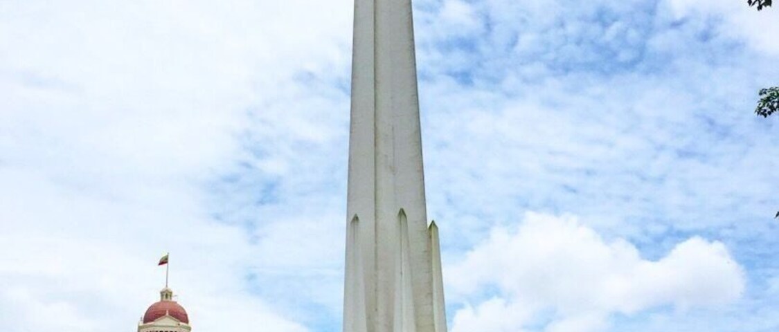 Un monumento alto en un parque, con una torre del reloj al fondo.