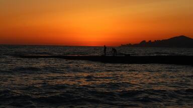 A sunset over the ocean with silhouettes of people on a pier.