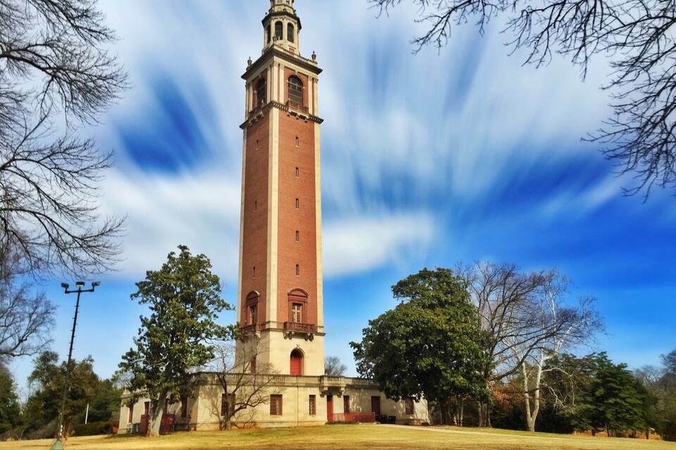 A tall brick tower with a dome and clock, surrounded by trees and a clear sky.