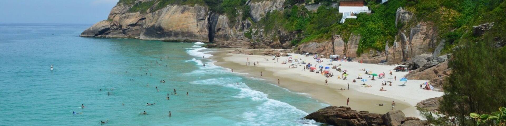 A coastal beach with people swimming and sunbathing, surrounded by lush greenery and buildings on a hillside.