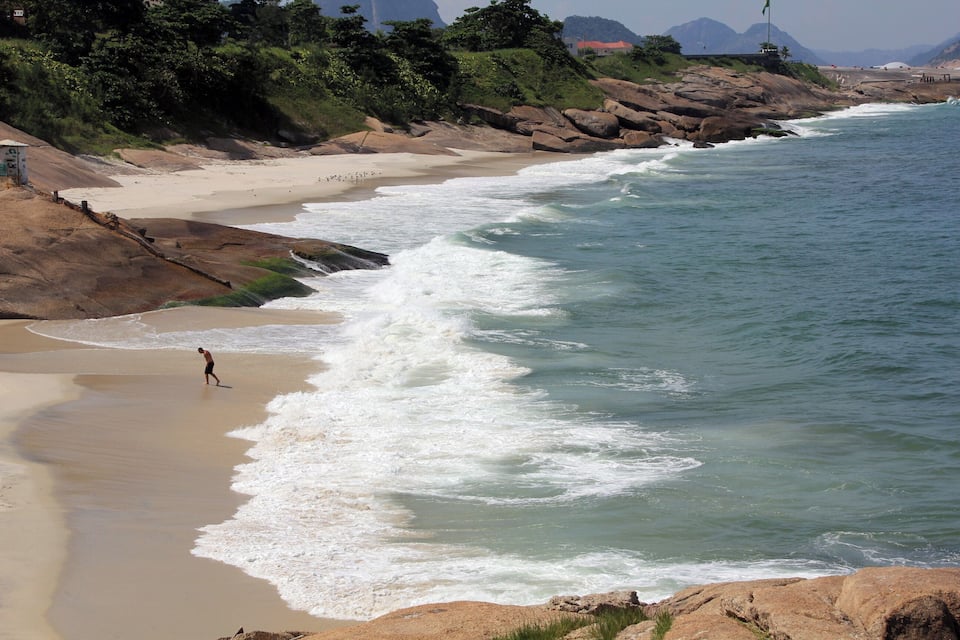 A sandy beach with waves crashing onto the shore, a person walking along the water's edge, and a rocky coastline with greenery in the background.