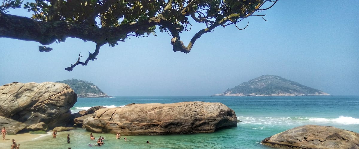 A rocky beach with clear turquoise water, people swimming and sunbathing, and distant mountains.