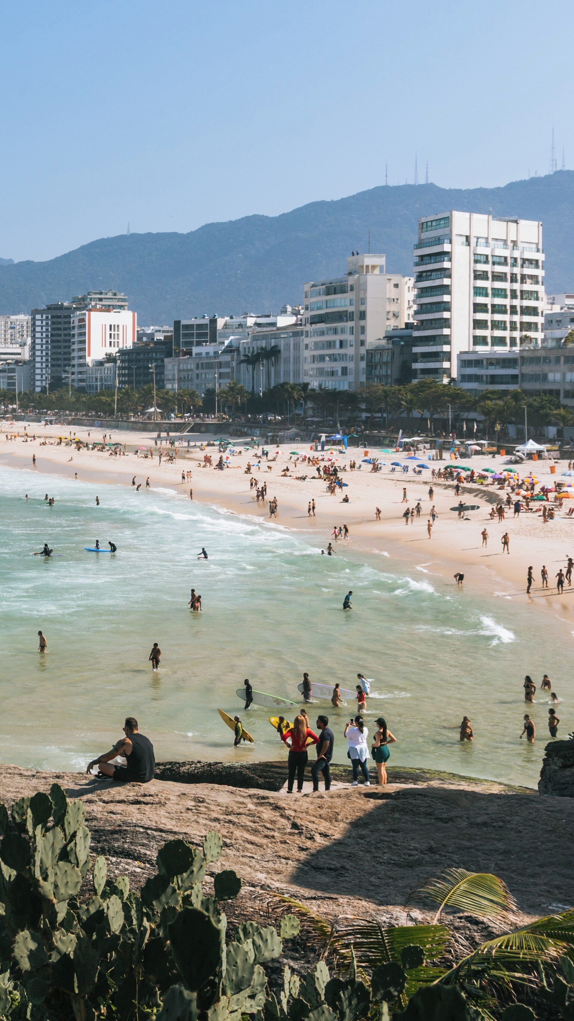 Visit to Pedra do Arpoador in Ipanema showcases a vibrant beach scene with surfers, sunbathers, and stunning mountains in the background