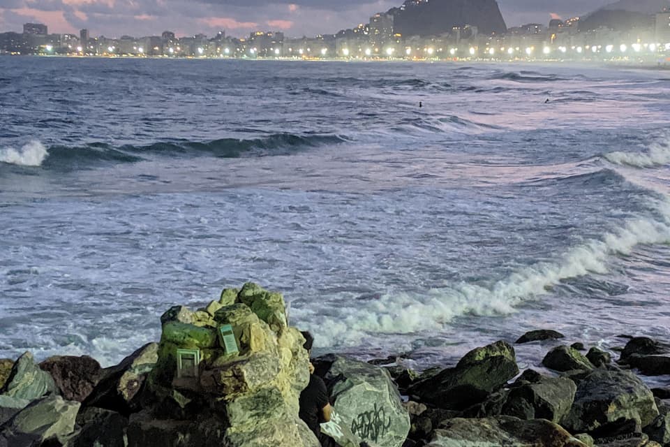 Un littoral rocheux où les vagues s’écrasent contre les rochers. Au loin, on aperçoit la silhouette d’une ville avec ses bâtiments et une montagne imposante.