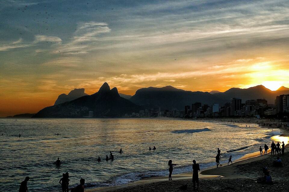 A beach with people swimming and walking, mountains in the background, and a sunset.