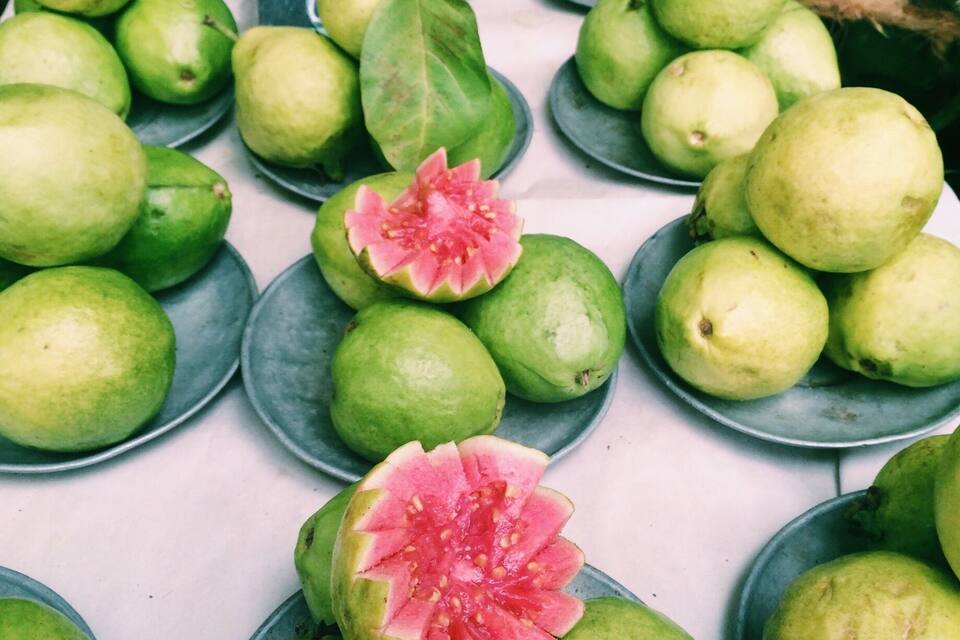 A market display of guavas, some cut open to reveal the pink interior, with a sign indicating 'Goaba Mel Hermelha'.