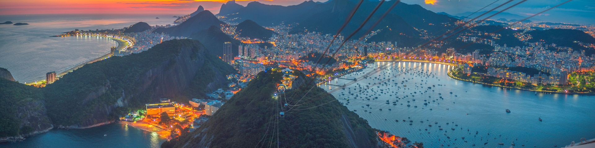 Vista panorâmica de uma cidade costeira com um sistema de teleférico, montanhas e uma baía cheia de barcos.