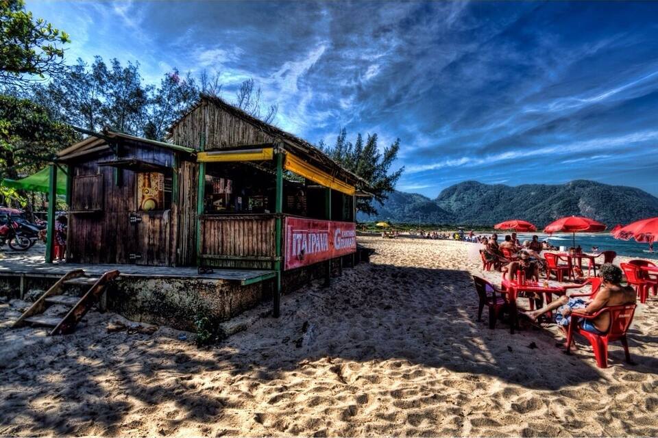 A beachside wooden shack with a red sign, surrounded by sand and trees, with people relaxing under red umbrellas.