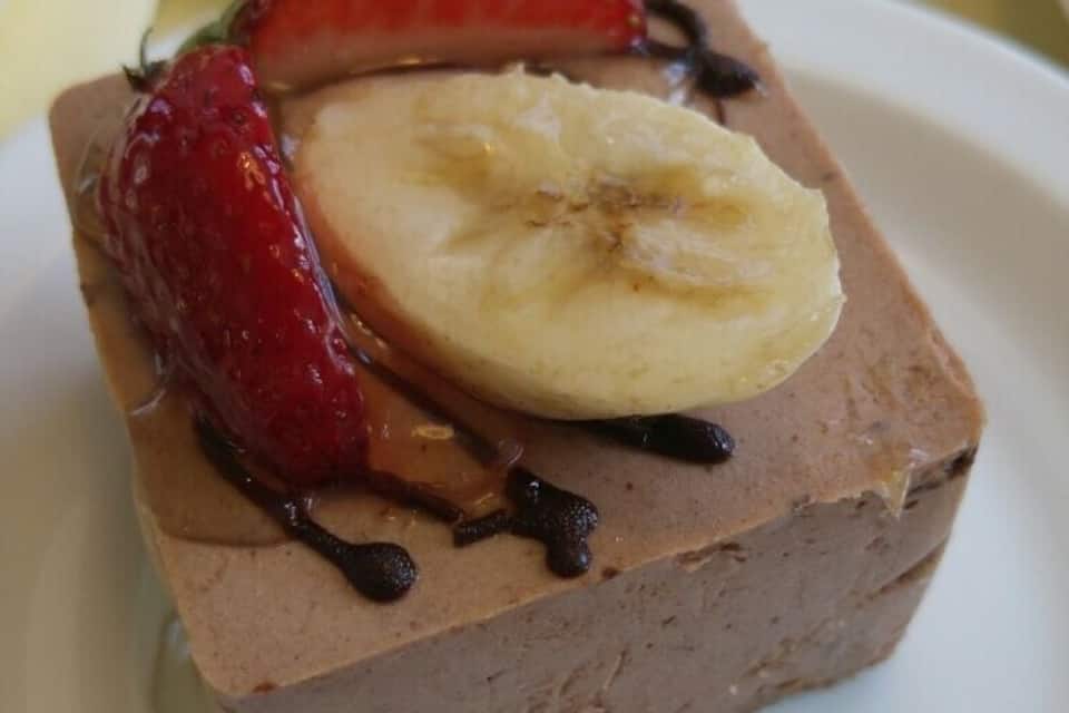 Un postre con mousse de chocolate, fresas, rodajas de plátano y un chorrito de chocolate sobre un plato blanco.