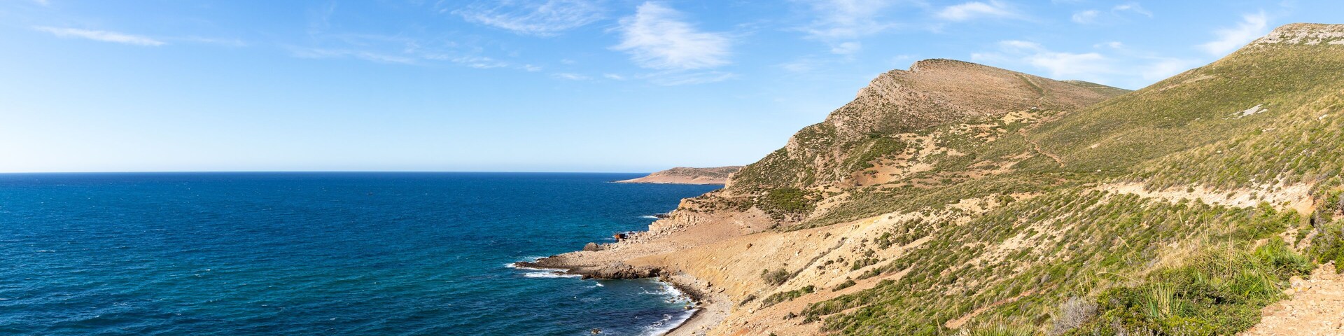 A coastal landscape with a clear blue sea, a sandy beach, and a rocky hillside with sparse vegetation.