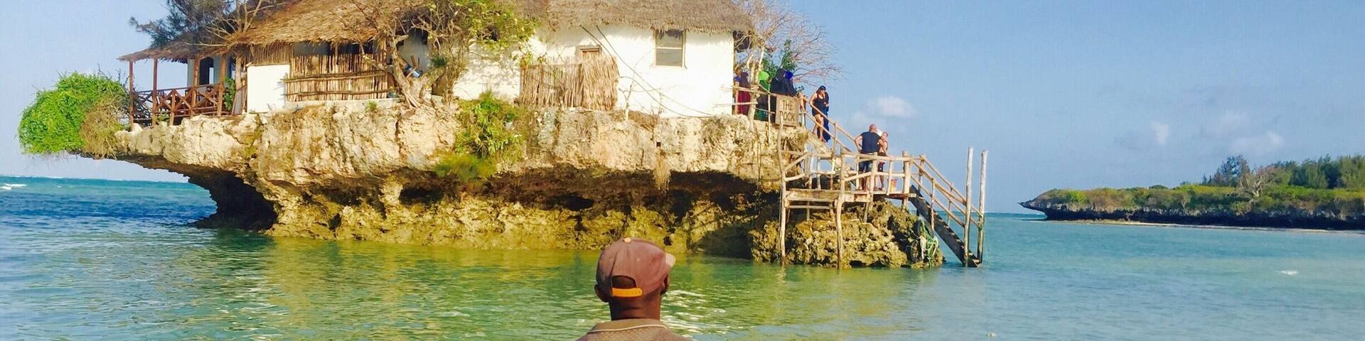 A man in a boat approaching a thatched-roof house on a rocky outcrop by the sea.