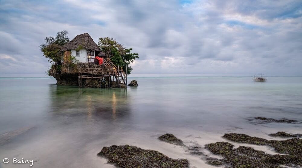 A thatched-roof house on stilts over calm sea water, surrounded by rocks and vegetation.
