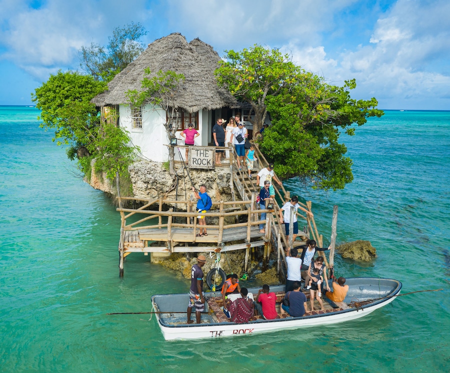 Eine Hütte mit einem Reetdach auf einer kleinen Insel, umgeben von türkisfarbenem Wasser.