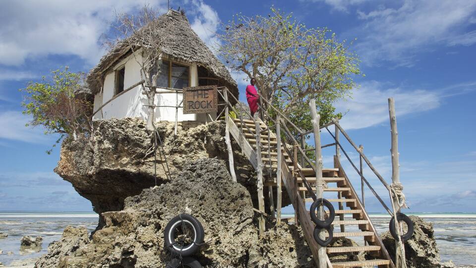 A thatched-roof hut on a rocky outcrop with a wooden staircase and a sign reading "THE ROCK".