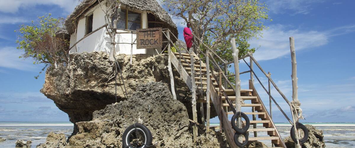 A thatched-roof hut on a rocky outcrop with a wooden staircase and a sign reading "THE ROCK".