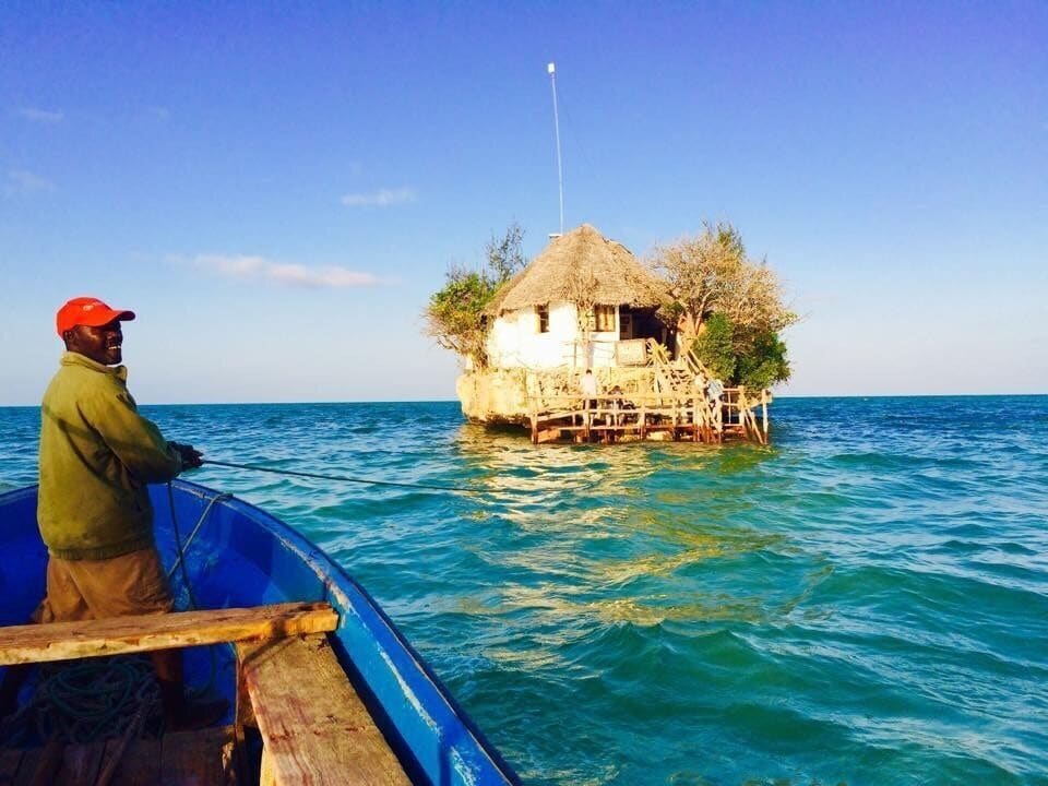 A person in a boat approaching a thatched-roof house on stilts in the water.