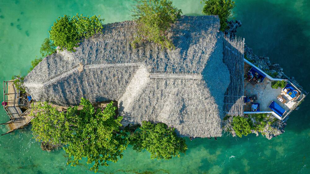 An aerial view of a small island with a thatched-roof structure, clear turquoise waters, and lush greenery.