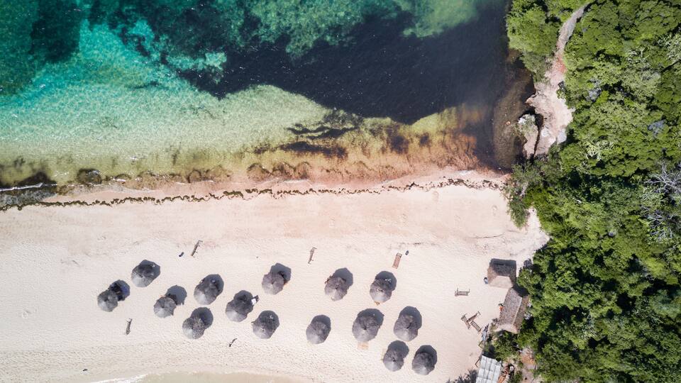 An aerial view of a beach with clear turquoise waters, a sandy shore, and a row of umbrellas.