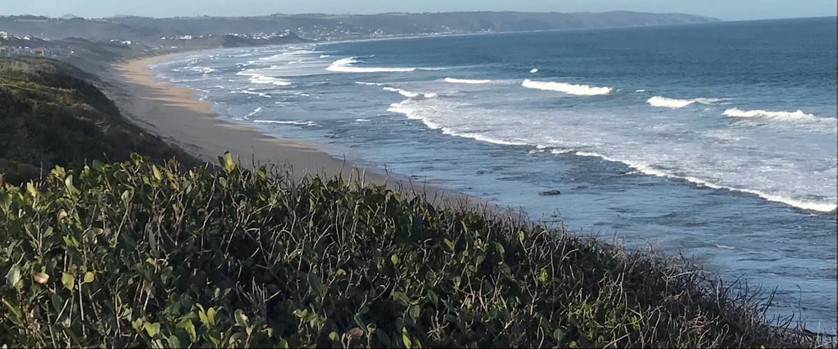 A coastal landscape with a sandy beach, ocean waves, and distant hills.